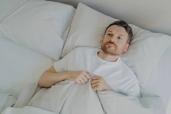 View from above of lonely upset young bearded man lying in bed with open eyes and can\'t sleep, feeling unhappy and tired, squeezes blanket with hands and being stressed of insomnia. Sleeping problems