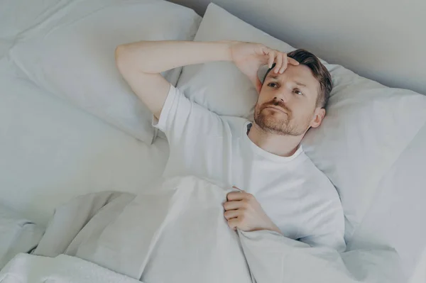 Young upset and tired bearded man lying on bed with open eyes and cannot sleep, male having insomnia problem or sleeping troubles. Bedtime and rest difficulty concept