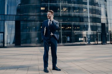 Koyu mavi takım elbiseli yakışıklı, genç, sakallı bir ofis çalışanı iş merkezinin yanında partnerlerin buluşmasını beklerken cep telefonuyla konuşuyor. İş adamları konsepti