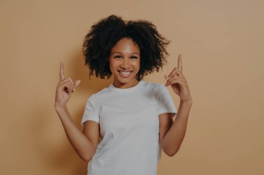 Genç, neşeli, kıvırcık saçlı, beyaz tişörtlü, işaret parmaklarıyla işaret eden, reklam için boş fotokopi alanı gösteren, bej renkli stüdyo arka planında izole edilmiş, heyecanlı, esmer bir kadın.