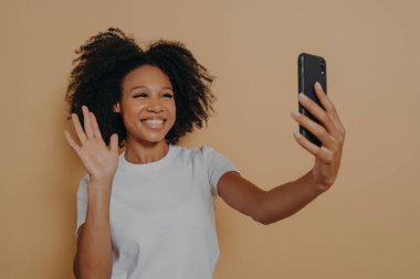 Hello! Beautiful cheerful dark skinned woman smiling at camera on modern smartphone, taking selfie on mobile phone and feeling happy, gesturing hi while making video call, isolated over studio wall