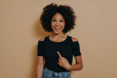 Young mean me? Portrait of happy young mixed race woman pointing finger at herself and looking at camera with amazed face expression, dressed in black t shirt. Body language and human emotions