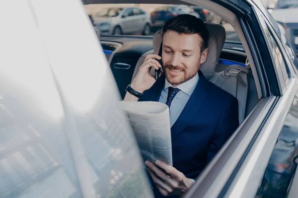 Good looking happy executive manager in stylish formal wear reads newspaper on backseat of car in same time speaking on smartphone with business partner, while staying in road traffic