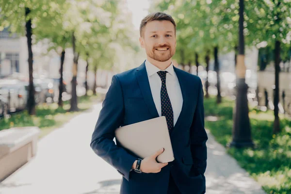 Cheerful young office worker in formal suit happy with end of successful corporate business conference walking along city park in sunny weather to meet new financial partner, with laptop under his arm
