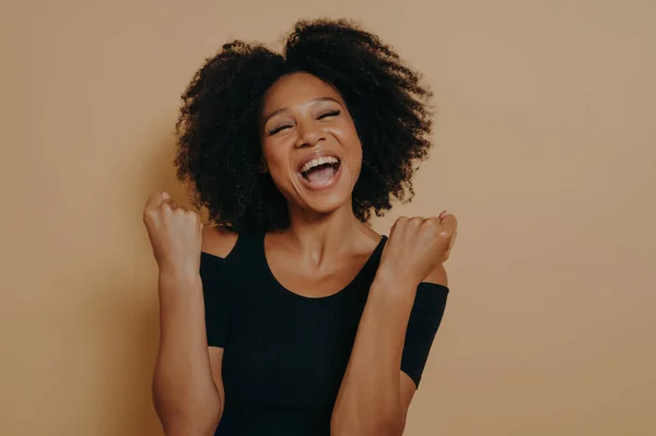 Studio shot of young joyful excited dark skinned woman keeping fists clenched and eyes closed, screaming from enjoyment while posing against beige background. Success and triumph concept