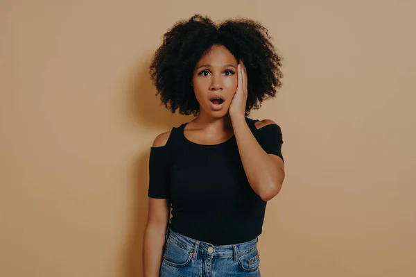 Studio portrait of young shocked african female with unexpected facial expression because of hearing bad news, mixed race woman keeping jaw dropped, wears casual outfit, posing against beige wall