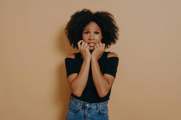 Woman about to panic hearing shocking worried news. Portrait of intense nervous and anxious dark-skinned female clenching fists opening mouth looking concerned over dark beige wall with copy space