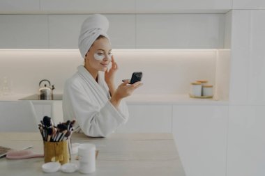 Kafasında bornoz ve havlu sarığı olan genç çekici bir kadın siyah halkaların altında kozmetik bantlar uyguluyor, kompakt aynaya bakıyor ve evde modern mutfakta dururken gülümsüyor.