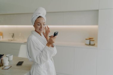 Kendine çok iyi bak. Bornozlu genç bir kadın aynaya bakıp gülümsüyor ve modern mutfakta dururken cilt bakım ürünleri uyguluyor ve evde estetik ameliyatlar yapıyor.