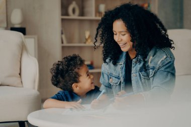 Uzun siyah kıvırcık saçlı mutlu, melez anne portresi. Küçük oğluyla oturma odasında oturmuş birlikte vakit geçirip resim yapmayı öğreniyorlar. Doğum günün kutlu olsun
