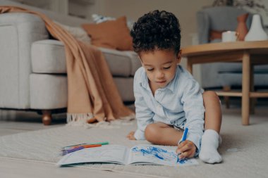 Küçük odaklı Afrikalı bebek oturma odasında halı döşenmiş bir zeminde oturuyor, boyama kitabında mavi kalemle çiziyor. Çocukların evde boş zamanları ve çocuk konsepti için yaratıcı hobiler.