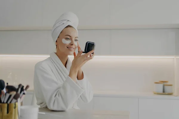 Morning skin care routine. Young beautiful woman in bathrobe and hair wrapped in towel applying cosmetic patches under eyes from dark circles and looking in compact mirror while standing in kitchen