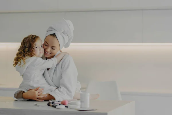 Young family mother and cute little daughter in white bathrobes hugging after spa procedures while standing in white modern kitchen at home. Beautiful loving mom spending time with child in morning