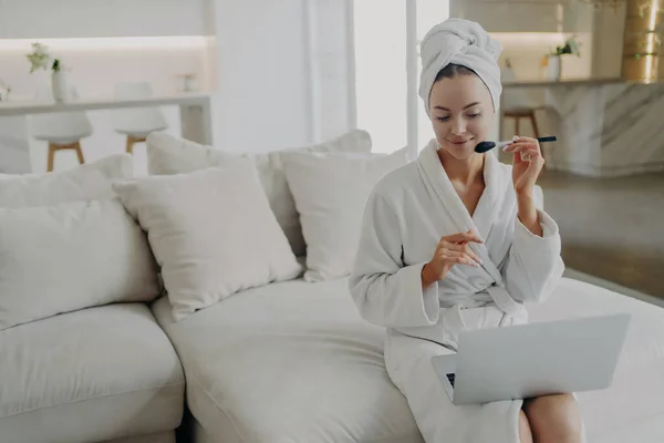 Young attractive woman in bathrobe and towel on head applying powder on face while watching make up video tutorial on laptop, sitting on sofa in modern living room at home and getting ready for work