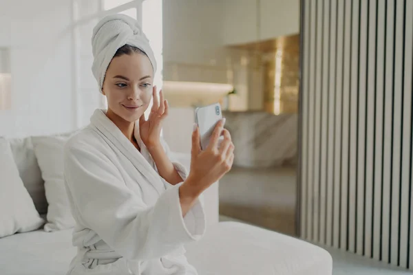 Beauty routine. Young gorgeous woman touching her soft healthy skin, applying cosmetic cream while sitting on sofa, female wearing bathrobe and towel on head holding smartphone in front of her face