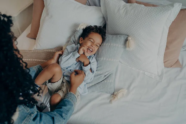 View from above of young loving afro american mother with long black curly hair tickling her cute little dear son before changing his clothes after coming home, mom and kid having fun on bed