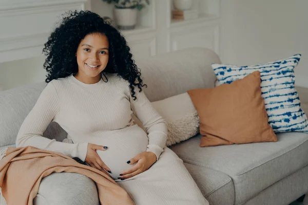 Happy pregnancy time. Cheerful young expectant lovely mixed race young mother with natural looking curly hair relaxing on sofa in living room, touching belly and smiling on camera