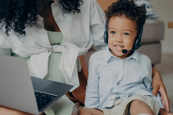 Happy afro american child with curly hair looking aside and smiling, wearing headset while having online meeting with dad, using laptop with his mother, boy talking with family through webcam