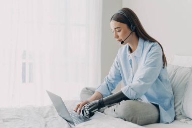 Handicapped freelancer has online meeting sitting on her bed. Young caucasian woman with artificial arm is working on laptop in bedroom. Girl is using headphones and microphone and chatting.