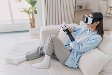 Attractive disabled girl is relaxing in vr glasses on couch. Handicapped caucasian person gets rehabilitation for sensor cyber arm. Amputee is setting functions of myoelectric robotic limb.