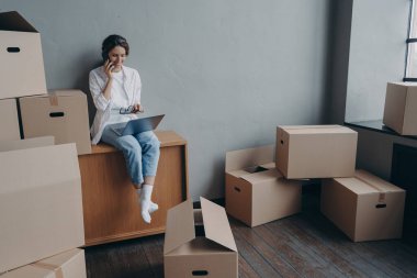 Happy homeowner talking on phone. Hispanic girl is sitting among carton boxes and working on pc. Attractive young business woman moves. Mortgage loan and real estate purchase.