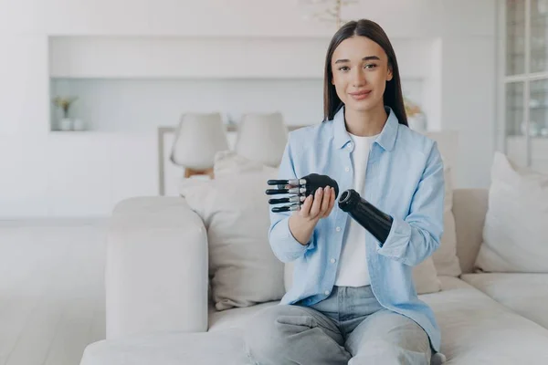 Positive disabled young woman sitting and disassembling bionic arm prosthesis. Cyber sensor artificial arm has processor chip and buttons. European girl has high tech carbon robotic hand and smiling.
