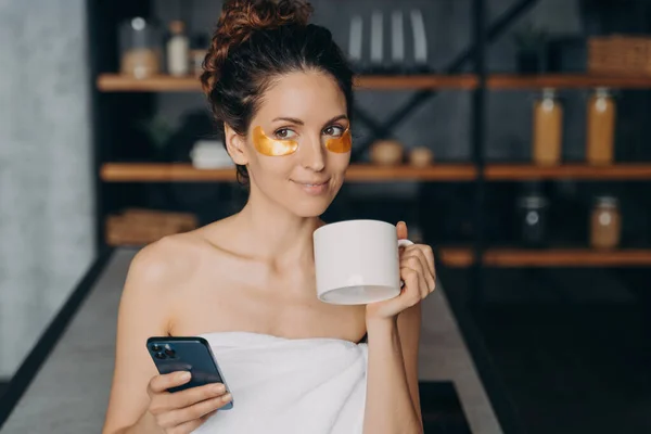 European girl drinks coffee and texting on smartphone. Young woman applies anti wrinkle eye patches and relaxing. Happy hispanic girl wrapped in towel after bathing. Evening body care routine.