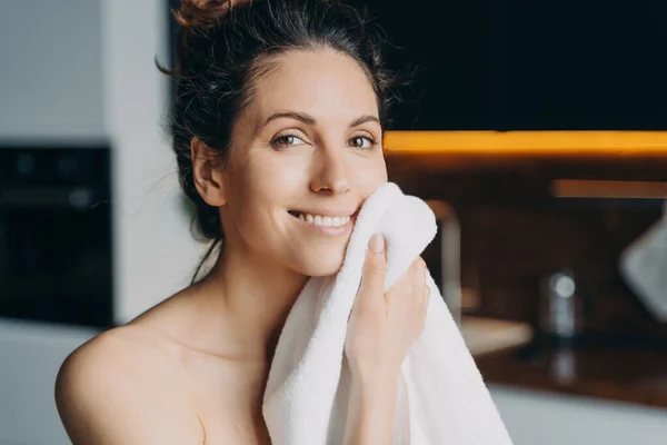 European girl is wiping face with towel after washing. Young brunette woman takes shower at home and doing skin care. Hygiene and freshness, dermatology and spa procedures.
