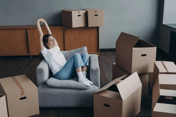 Lady is relaxing among boxes in living room of new home. Happy woman is an apartment buyer. Satisfied european girl is sitting in armchair. Dream house purchase and mortgage concept.