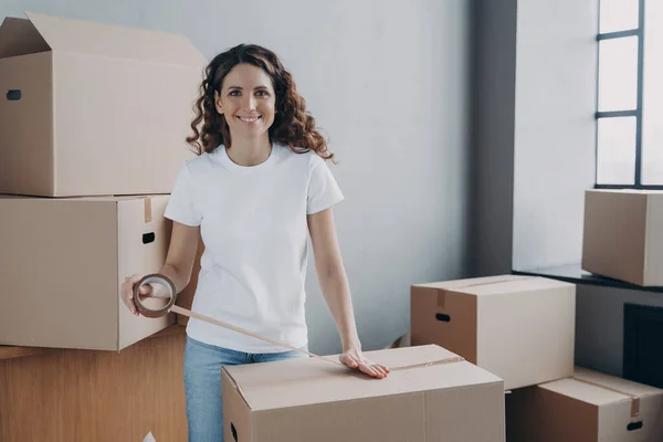 Young woman is loading and packing boxes for moving. European lady is wrapping cardboard boxes with packing tape. Moving service worker preparing boxes for shipping and storage. Delivery service.