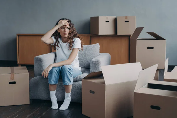 Headache from moving. Exhausted woman sitting among packed cardboard boxes. Spanish young lady in jeans and white t-shirt going to relocate. Depression and stress from moving concept.