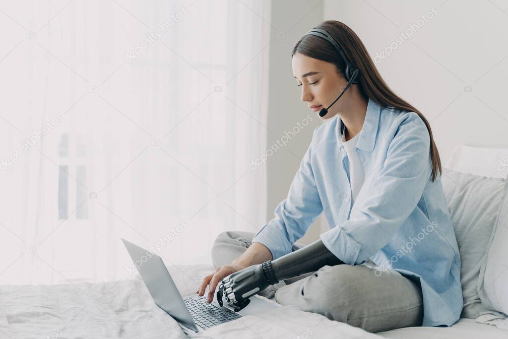 Handicapped freelancer has online meeting sitting on her bed. Young caucasian woman with artificial arm is working on laptop in bedroom. Girl is using headphones and microphone and chatting.