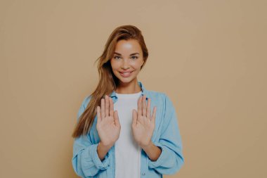 Parlak uzun saçlı, büyük mavi tişörtlü, beyaz tişörtlü, gülümseyen genç bayan model stüdyoda bej arka planda dururken dur işaretini göstermek için elleri kullanıyor. Olumlu kadın duyguları