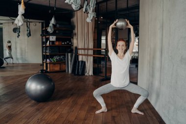 Fiziki olarak iyi durumda, spor yapan bir kadın spor salonunda barmenlik yapıyor, Plie 'de oturuyor ve iki eli de başının üstünde dizlerinin üzerinde küçük bir fitball topu tutuyor.