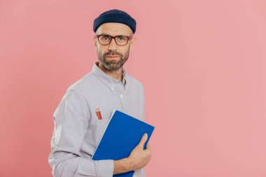 Yakışıklı erkek Tasarımcısı şık giysiler giyiyor, gömlek cebinde iki kalem vardır, skeçler, promosyon metninizi için boş alan ile pembe arka plan üzerinde izole yapmak için mavi Not defteri tutar