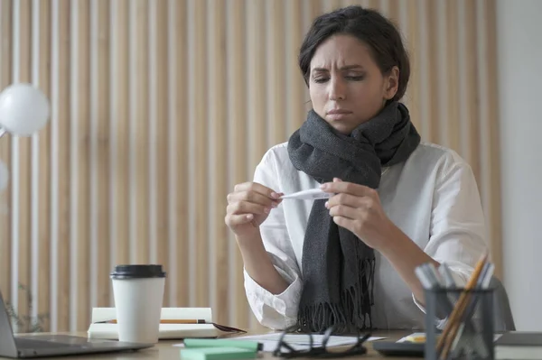 Unhealthy young hispanic business woman wearing scarf around neck holding thermometer, feeling unwell while working in office, female employee sitting at table and suffering of high fever