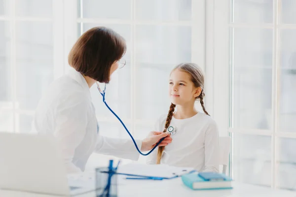 Woman pediatrician gives consultation to small girl, talk about health and symptoms, listens heart with phonendoscope, makes prescription, pose in hospital office. Childrean medical insurance and care