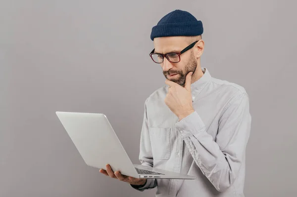 Attentive student holds chin, focused in monitor of laptop computer, searches information for project, connected to wireless internet, isolated over grey studio wall. Networking and people concept