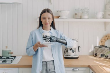 Modern caucasian disabled girl holding cup of coffee, using bionic prosthetic arm, standing in kitchen. Young woman holds mug by artificial hand at home. Lifestyle of people with disabilities concept.
