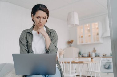 Thoughtful woman looking at laptop screen, pondering ideas, difficult working tasks at home. Pensive female touching chin, reading email, message, news in social networks. Remote job, elearning.