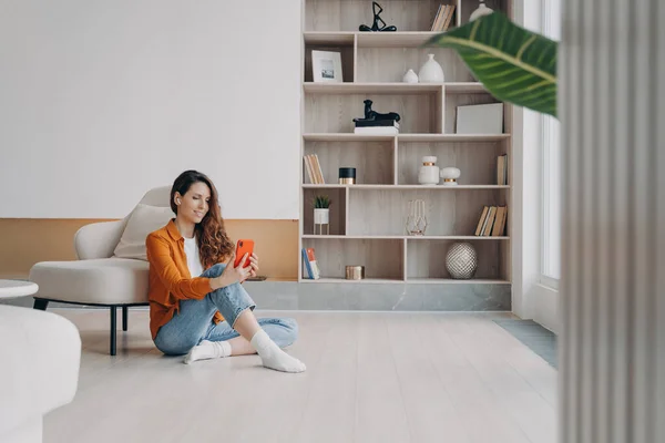 Smiling woman with wireless earbud using phone, looking at camera, sitting on heated floor in modern living room. Female having online video call or listening to music playlist or podcast at home.