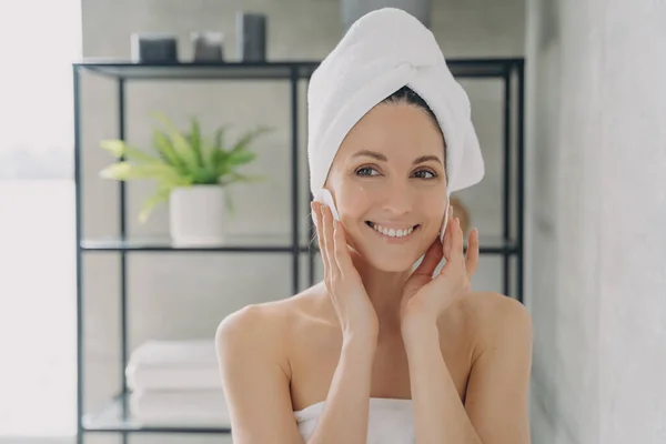 Female with towel on head cleanse face skin removing makeup using cotton pads after shower. Latina woman enjoying fresh clean healthy skin. Skincare beauty treatment, self care concept.