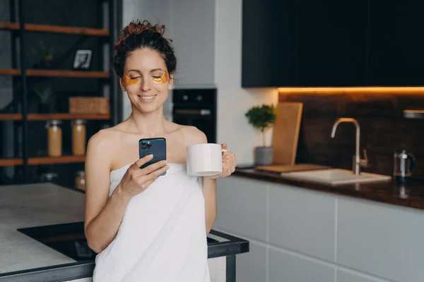 Modern young businesswoman with skincare under eye patches reads messages, news on smartphone, holding morning coffee cup. Happy girl wrapped in towel browsing social networks content after shower.