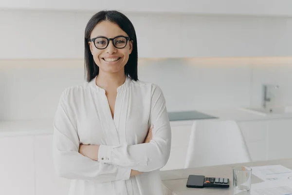 Young european executive or ceo in glasses and white blouse is working remote from home. Confident girl, entrepreneur is standing with her arms crossed and smiling. Career and leadership concept.