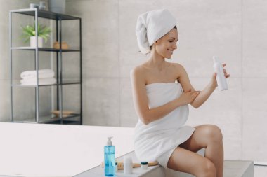 Smiling woman wrapped in towel applying organic body care lotion sitting on bathtub in modern bathroom. Female using sunscreen cream after shower. Self-care, skincare treatment, spa procedure