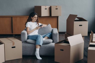 Female holding smartphone chooses relocation service or looking for repair ideas sitting in armchair with boxes. Smiling spanish woman tenant owner booking new dwelling on moving day.
