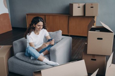 Smiling girl choosing mover online, using mobile phone app, sitting with cardboard boxes waiting to move to new home. Satisfied woman holding smartphone, arranging relocation, selects moving company.