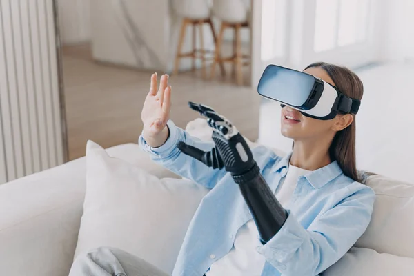 Happy disabled girl wearing virtual reality glasses playing in video game, training to use her bionic prosthetic arm, sitting on sofa at home. High tech for rehabilitation of people with disabilities