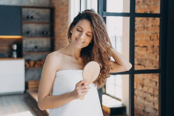 Pretty woman combing hair feels satisfied after beauty treatment procedure, smiling hispanic girl wrapped in towel after shower brushing curly long brown hairs at home. Haircare cosmetics advertising.