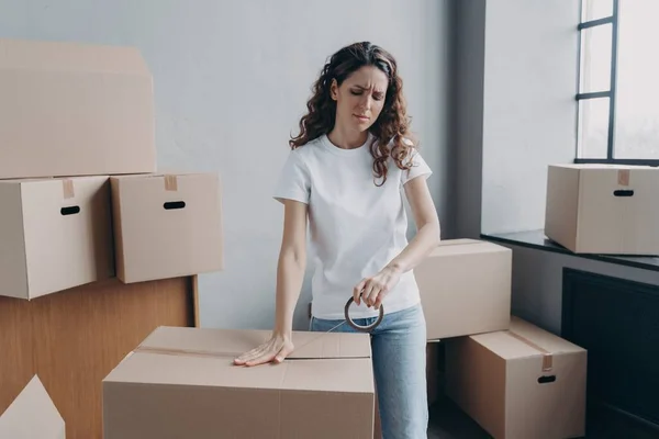 Tired hispanic woman packs cardboard boxes using duct tape. Overworked female employee packs parcels with goods with adhesive sellotape, frowning. Overtime, delivery service concept.